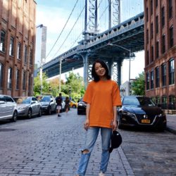A photo of our Student Sustainability Architect Freya Wang, who is wearing an orange top and blue jeans and is stood in front of a metal bridge.