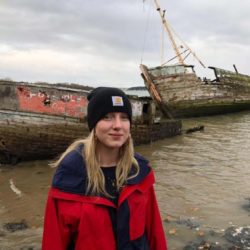 A photo of Imogen Lee-Savage, who is smiling in a red raincoat and black beanie in front of a shipwreck.