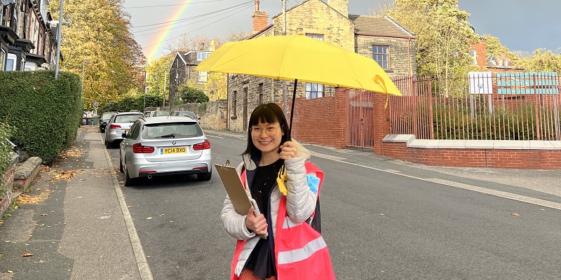 Photo of a woman wearing a pink high vis holding a yellow umbrella on a street with a rainbow in the background