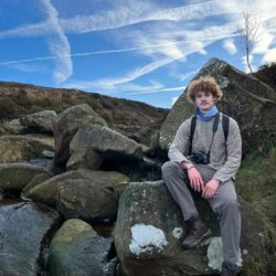 A photo of Sam Brooke sat on a boulder in the countryside, wearing grey walking clothes with a camera around his neck.