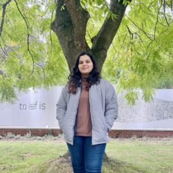 A photo of Student Sustainability Architect Rasika, who is smiling at the camera in front of the sculpture outside Ester Simpson