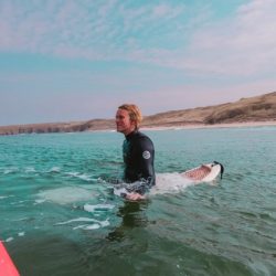A photo of Student Sustainability Architect Jamie sat on a surfboard in the sea, dressed in a wetsuit with wavy blonde hair