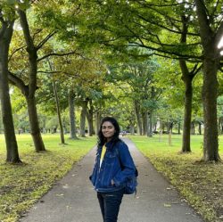 A photo of Student Sustainability Architect Spoorthi, who is wearing a denim jacket on the path of a park surrounded by trees