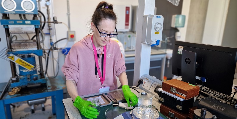 Photograph of Ashley in a pink jumper wearing green gloves stood at a bench in the lab