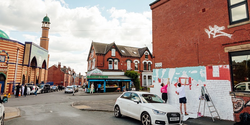 Photograph of two people painting a white and blue mural on the side of a red brick house in the leeds community