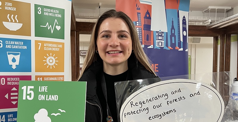A woman is smiling at the camera while holding a green SDG 15 'Life on Land' logo and a speech bubble with the words 'regenerating and protecting our forests and ecosystems' written on it.