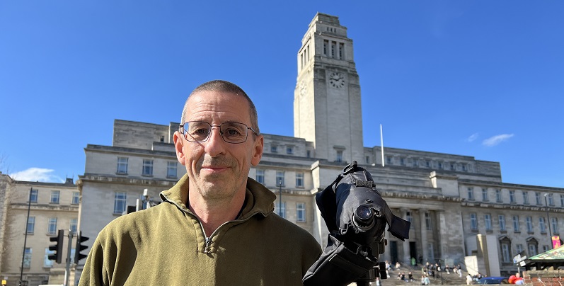 A photo of Les smiling in front of Parkinson building with his camera on a tripod