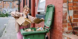 A photo of a woman with brunette hair putting some cardboard boxes into a green recycling bin outside a house, more information in the halls to homes 2023 document.