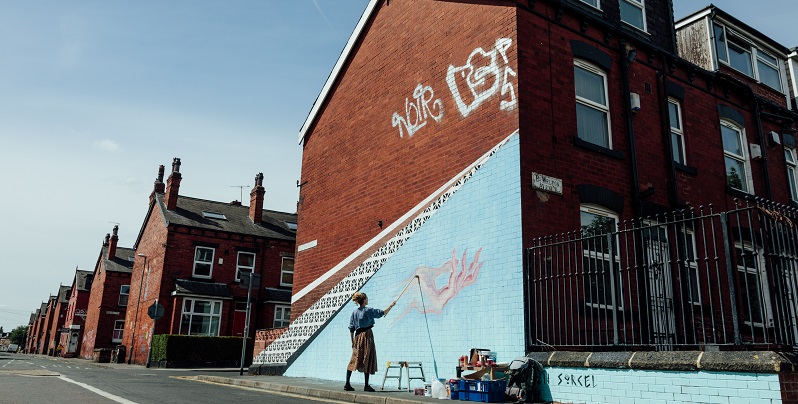 The side of a house in Leeds with a woman painting a large mural with a light blue background.