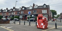 A red British Heart Foundation donation bank, in front of six black bollards in a terraced, red brick street.