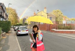 Woman stood smiling with an umbrella with a rainbow in the background.