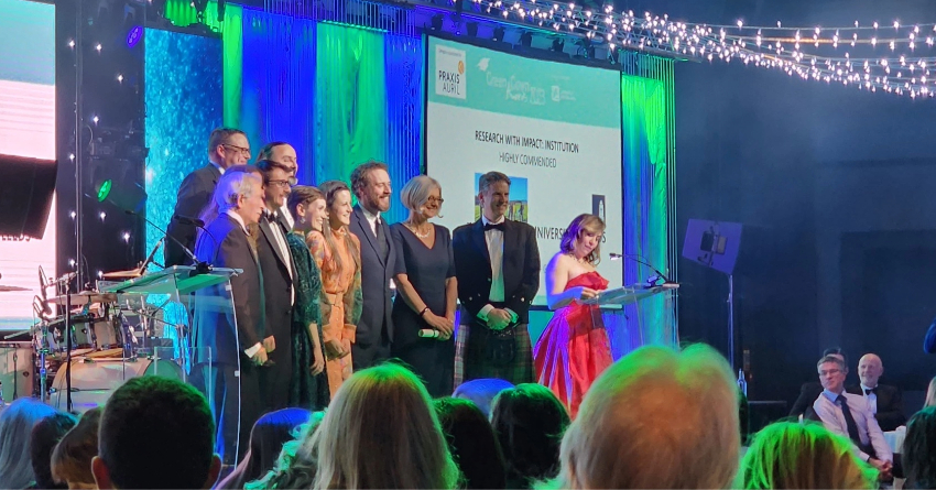 Picture shows a group from the University of Leeds on stage to receive an award. A lady wearing red is stood speaking to the audience.