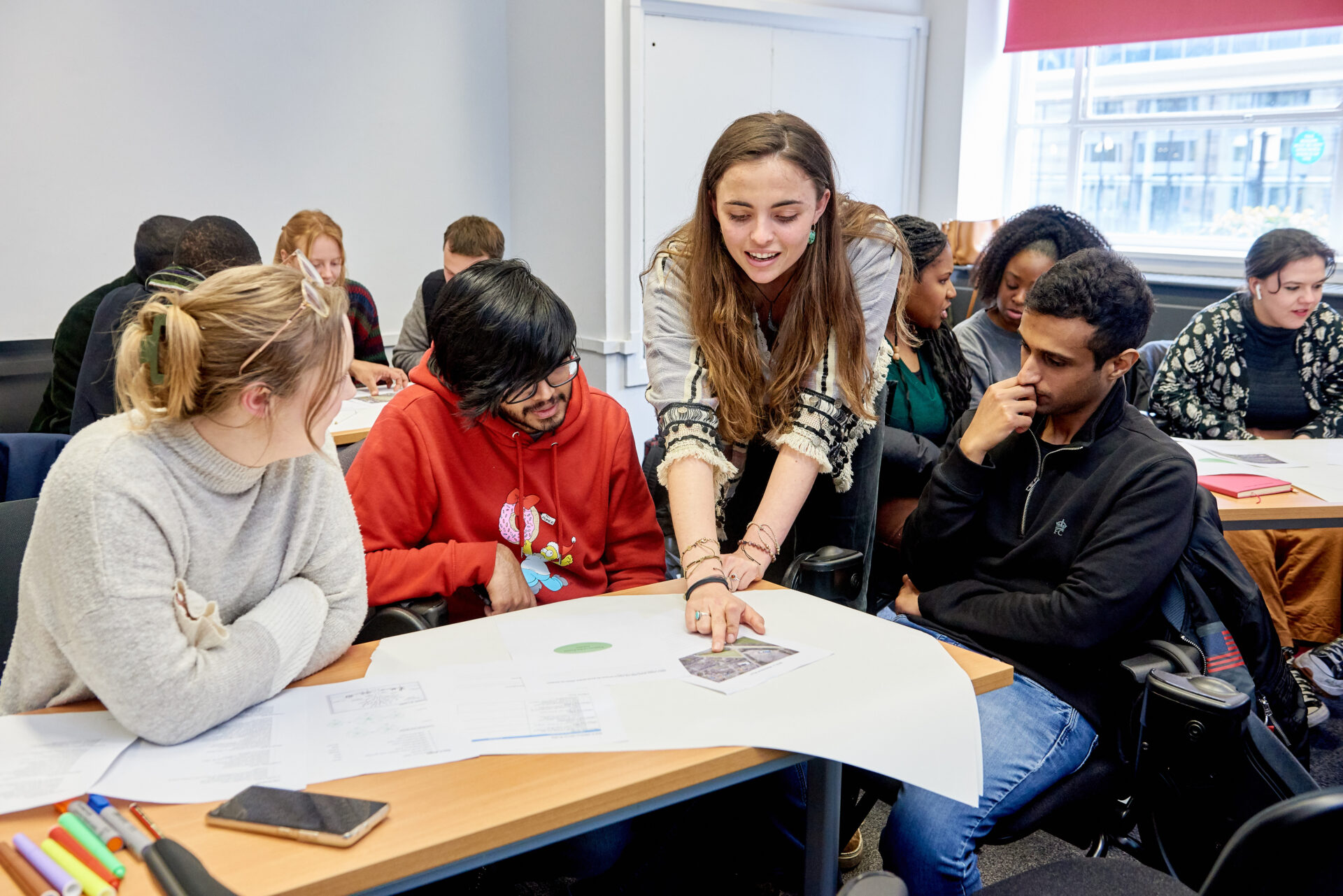 A student presenter points to a poster. A group of student conference attendees gather round