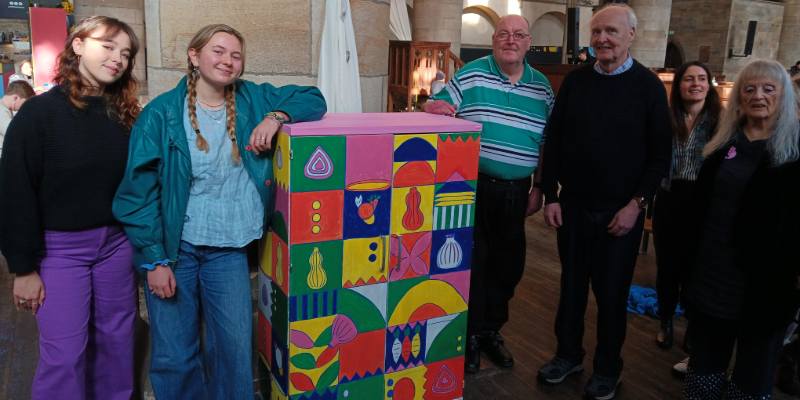 A group of people are stood around a brightly painted cupboard being used as a community pantry.