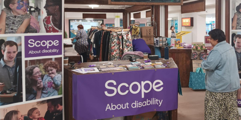 A stall in LUU by Scope. Clothes are hung on a rail in the background while at the front a banner and purple tablecloth show the name of the charity 'scope' and the words 'about disability'.