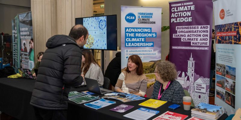 A man is talking to some people behind a table. There are Yorkshire and Humber Climate Commission and Climate Action Pledge banners behind them, and papers and leaflets on the table.