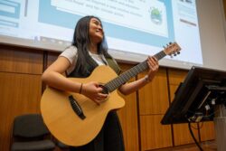 A student strums their guitar during their music performance at the Student Sustainability Research Conference. They have long dark hair and are smiling