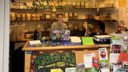 A person smiling behind the desk of the Green Action Co-op, where there are various food grains/ products in glass jars in the background.