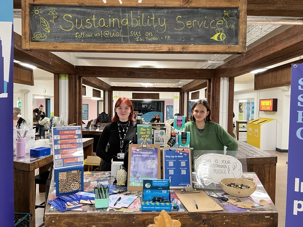 Two women stood behind a table with posters, pens and leaflets on it. There is a sign above the stall that says "Sustainability Service".