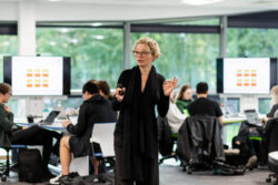 An academic tutor stood teaching in the middle of a large, busy classroom full of students. The modern room has big, bright windows along the side, with large digital screens and clusters of students working together on their laptops, around shared desks.